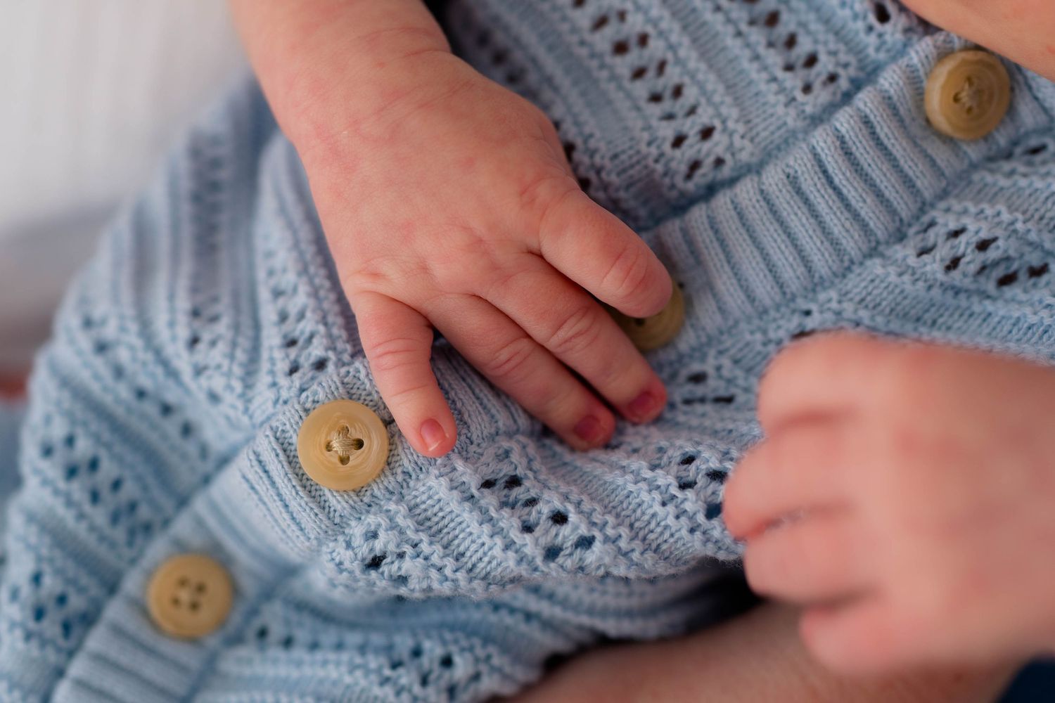 Close-up of newborn baby hands resting on a soft blue knitted romper with wooden buttons.