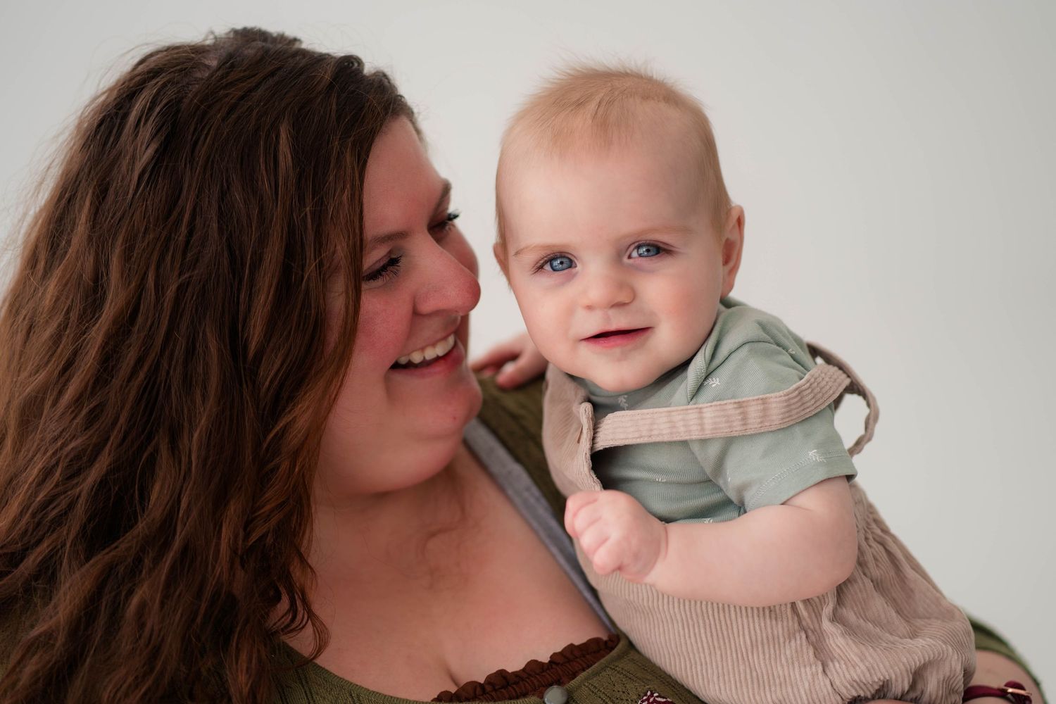 A smiling mother holds her happy blue-eyed baby wearing a green romper against a light background.