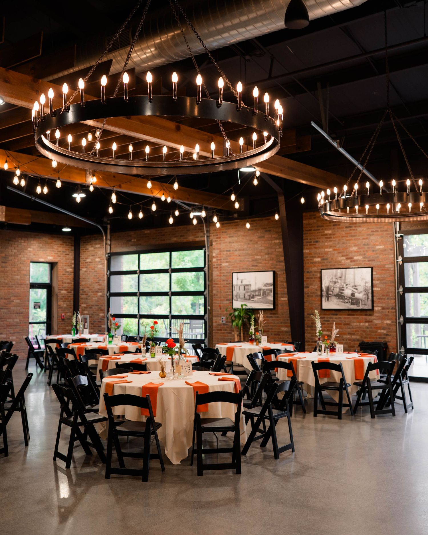 Elegant wedding reception hall with exposed brick walls, round chandeliers, and decorated tables with orange accents.