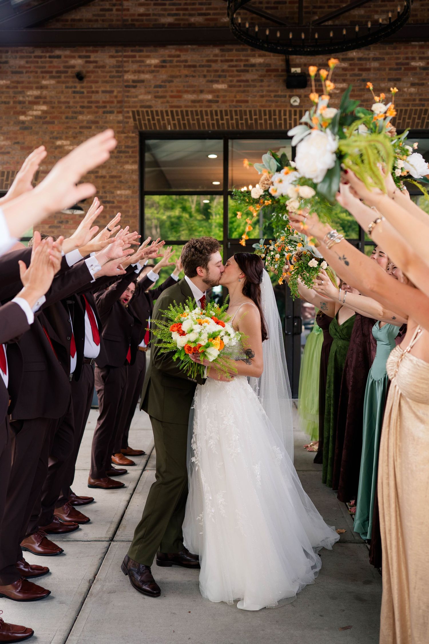 Newlyweds share a kiss during wedding party sendoff tunnel with raised arms and bouquets.