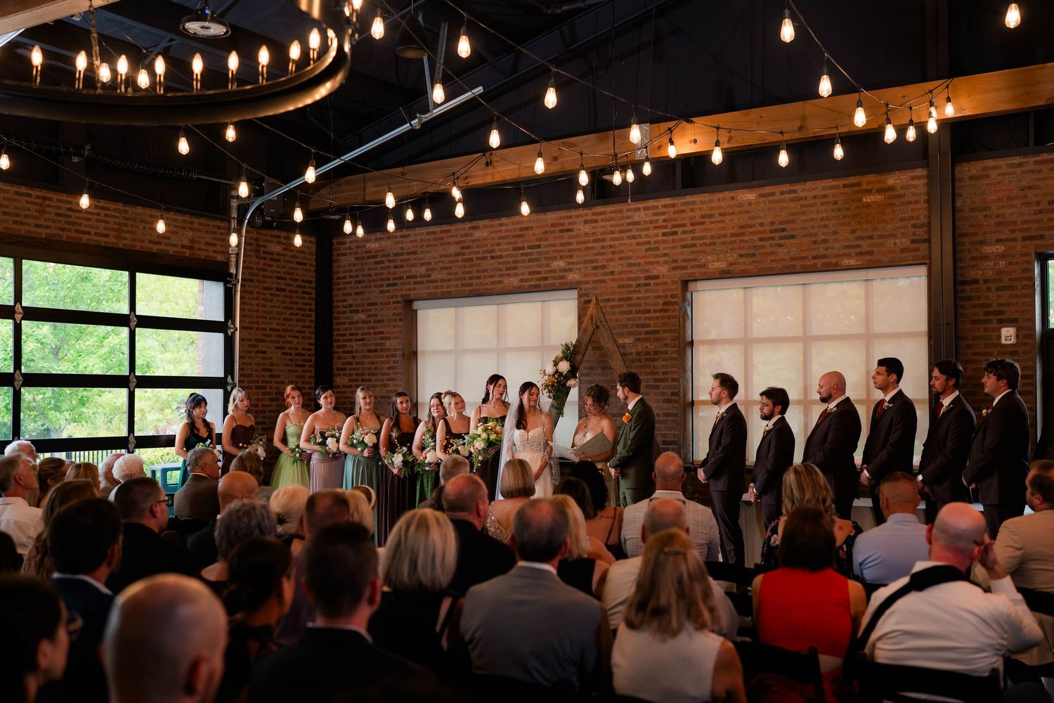 Wedding ceremony in industrial brick venue with string lights and full wedding party at altar.