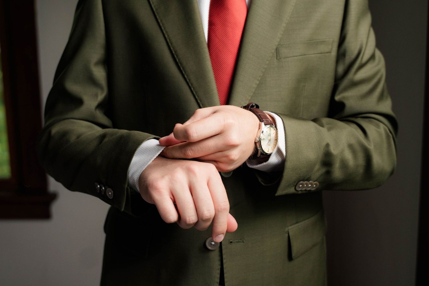 Man in olive green suit and red tie adjusting his brown leather watch on his wrist.
