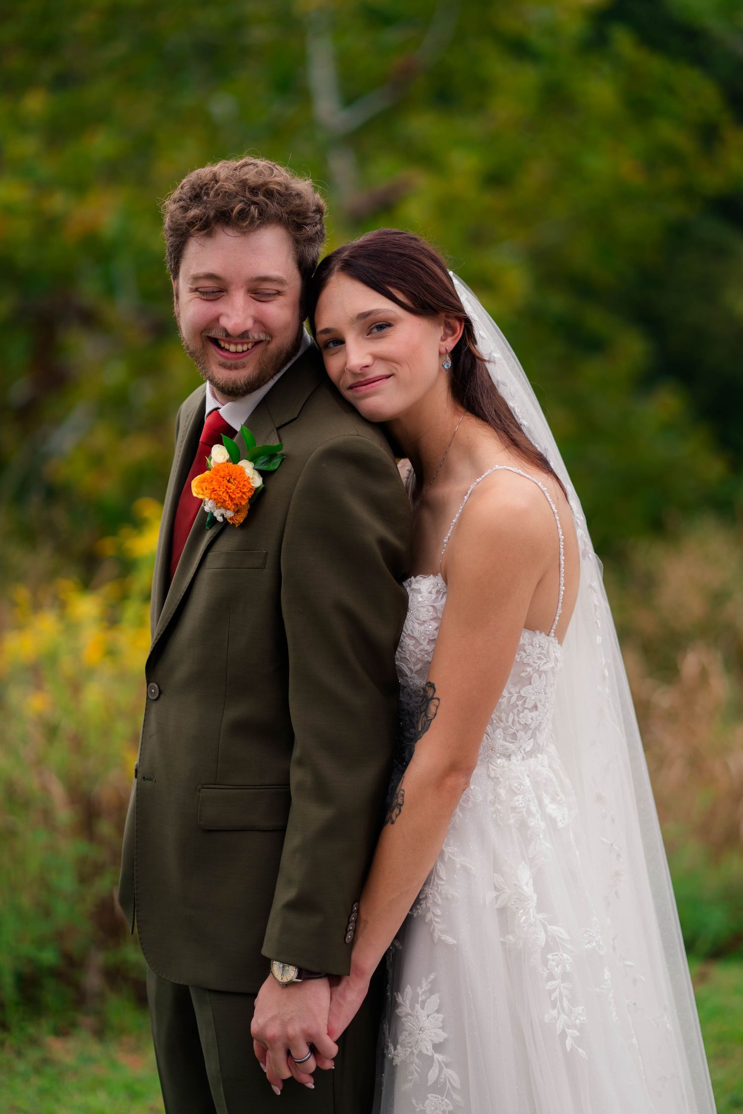 Smiling bride and groom holding hands outdoors with colorful autumn foliage background.