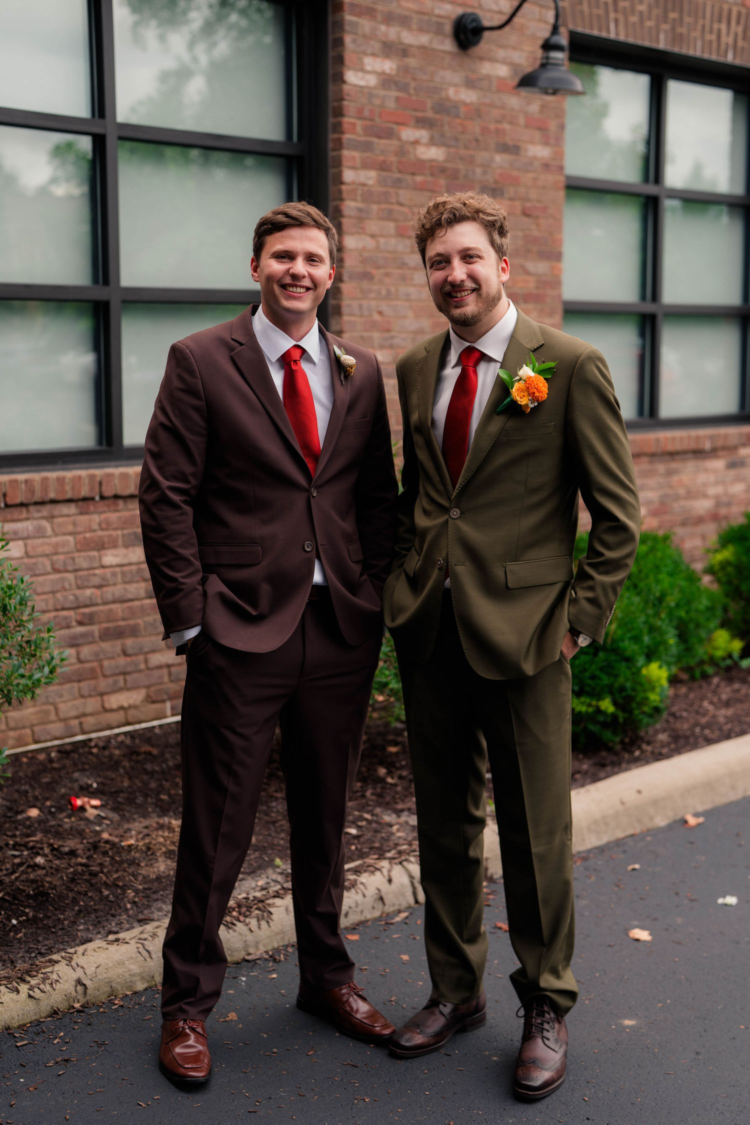 Two groomsmen in olive and brown suits with red ties and orange boutonnieres pose outside brick building.