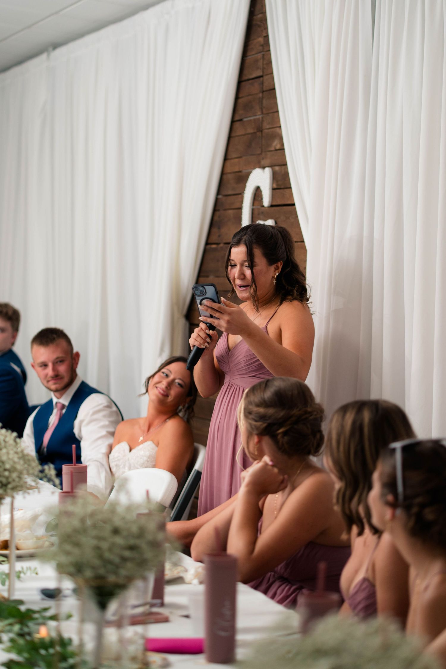 Bridesmaid reads from phone while giving a speech at wedding reception as bride laughs at head table.
