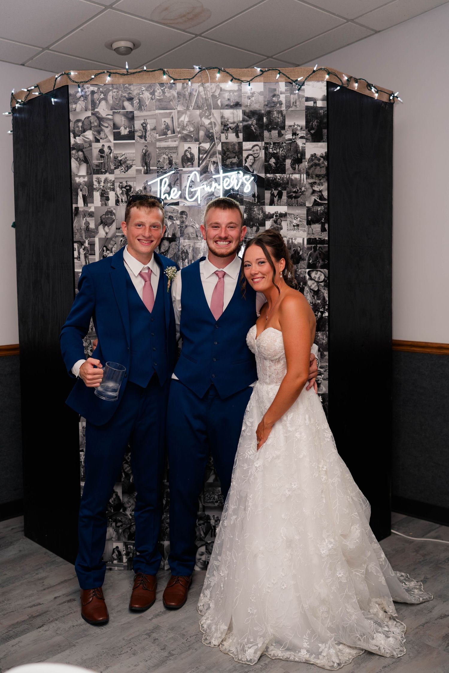 Bride and groom pose with groomsman in front of black and white photo collage backdrop at wedding reception.