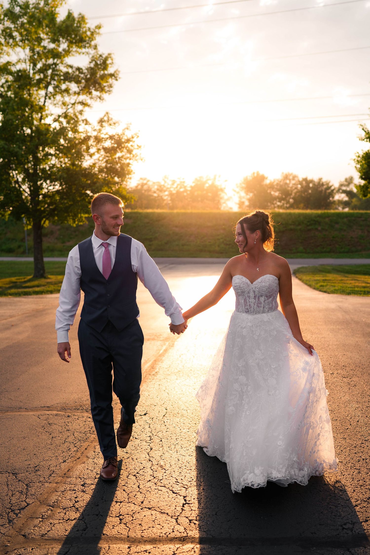 Bride and groom walk hand-in-hand down a road at golden hour, smiling at each other in wedding attire.