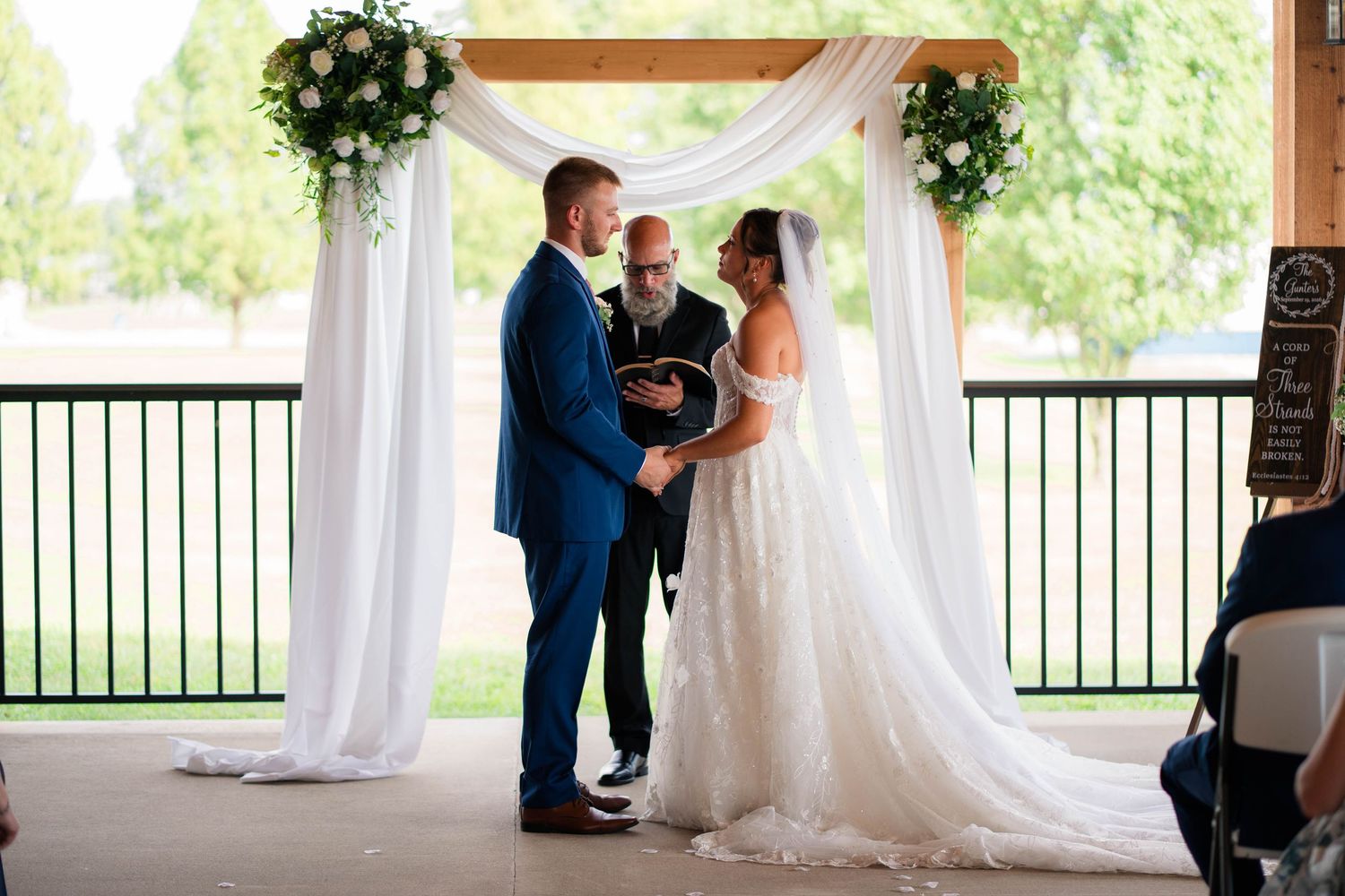 Bride and groom exchange vows under white draped wooden arch with floral arrangements.