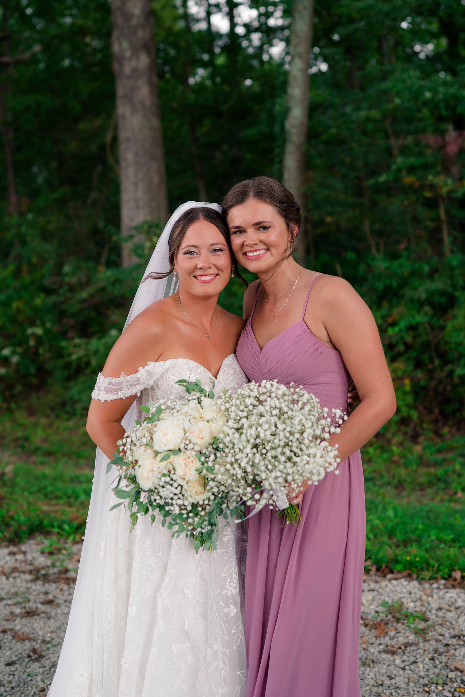 Bride in lace gown and bridesmaid in mauve dress holding bouquets of white roses and baby's breath outdoors.