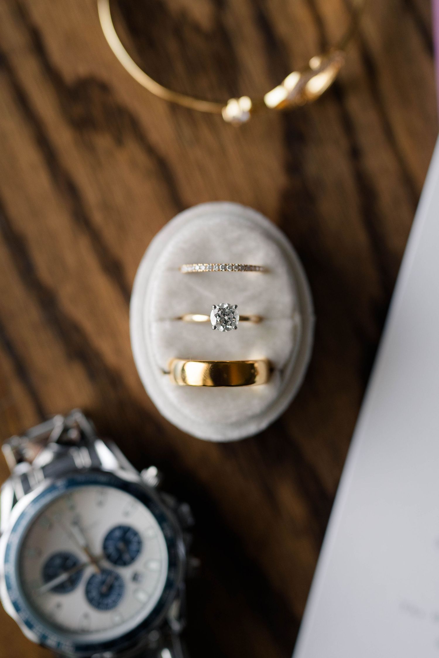 Gold diamond engagement ring and wedding bands displayed in oval velvet ring box with luxury watch.