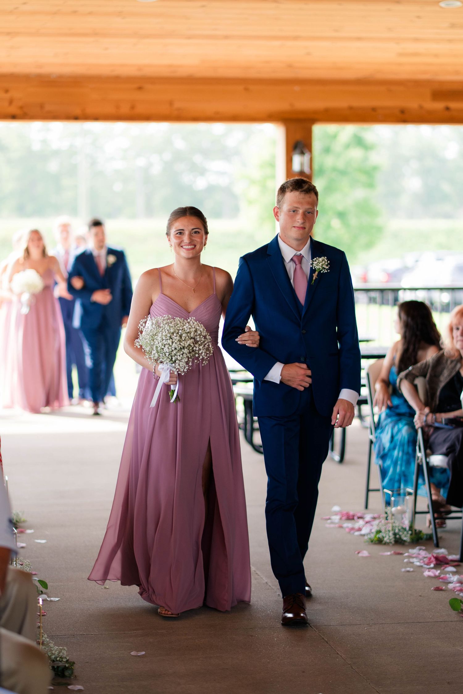 Bridesmaid in mauve dress holding baby's breath bouquet walks down aisle with groomsman in navy suit.