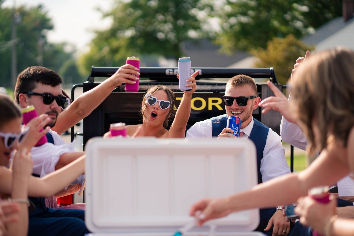 Wedding party celebrating on flatbed truck with drinks and sunglasses after ceremony.