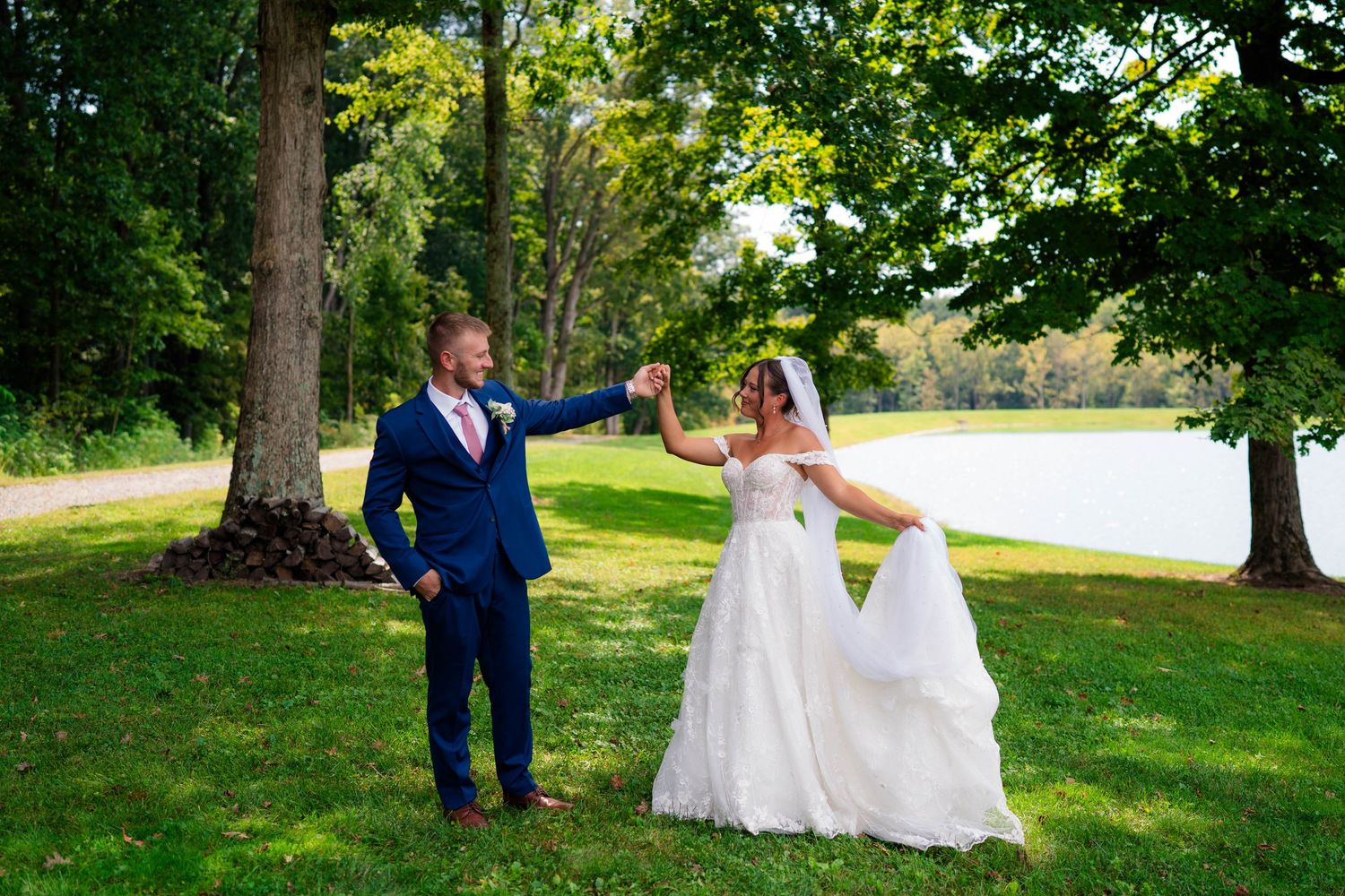 Groom twirling bride in white wedding dress on green lawn near lake surrounded by trees.
