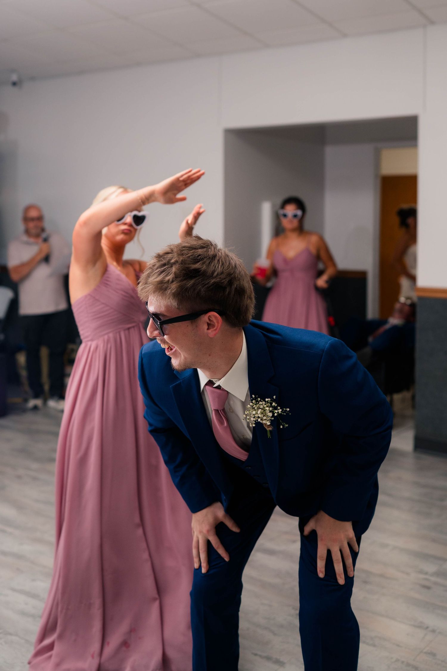 Groomsman in navy suit laughs while bridesmaid in pink dress dances behind him at reception.