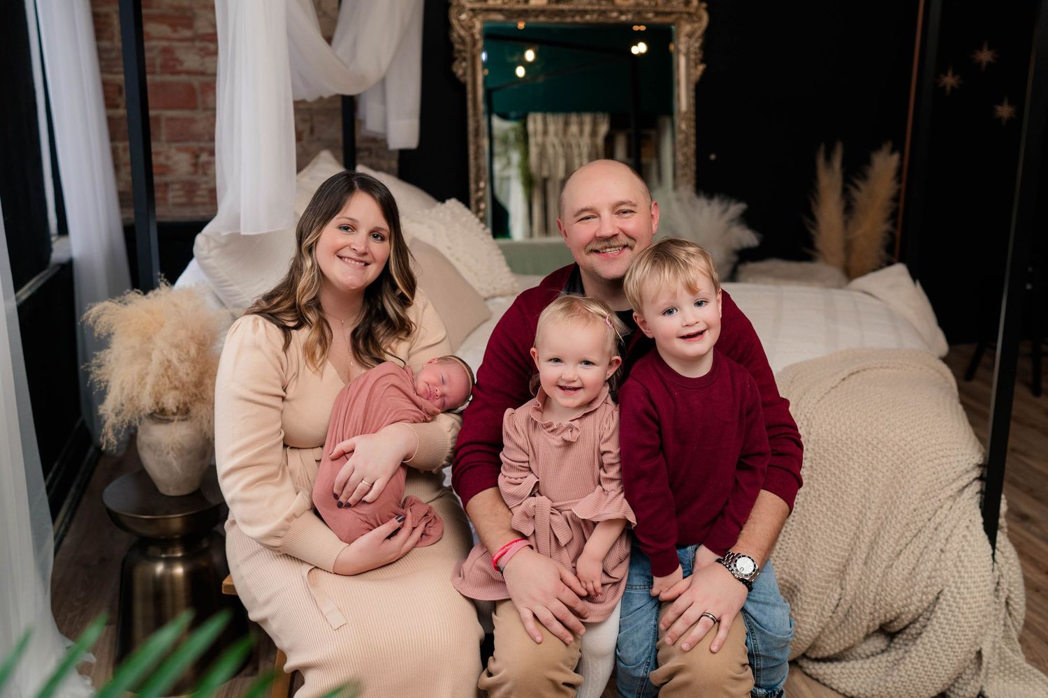 Happy family of five posing together on a bed, including parents, a toddler, young child, and newborn baby.