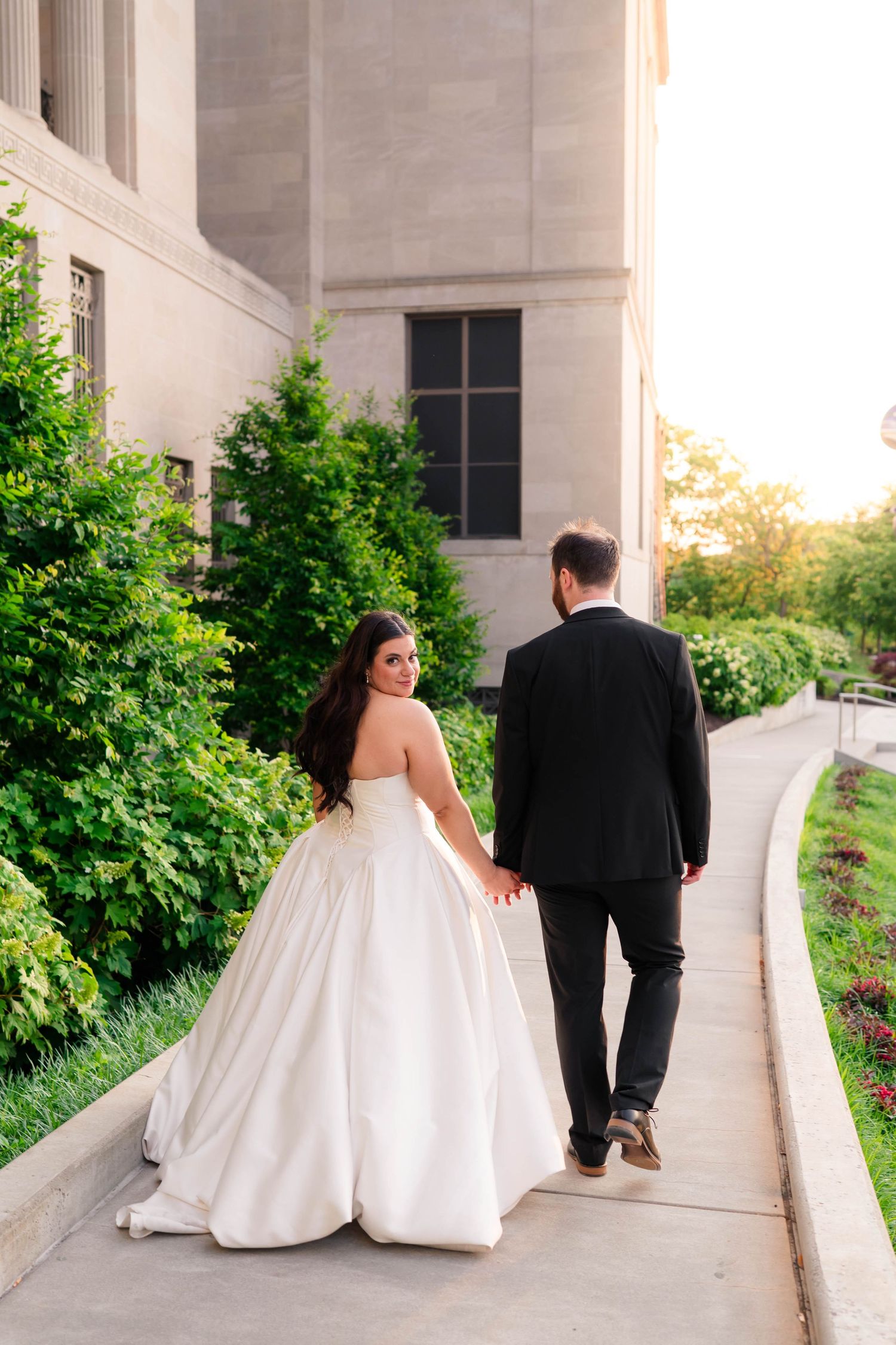 Bride looks back smiling as couple walks hand-in-hand along a garden path beside a grand stone building at sunset.