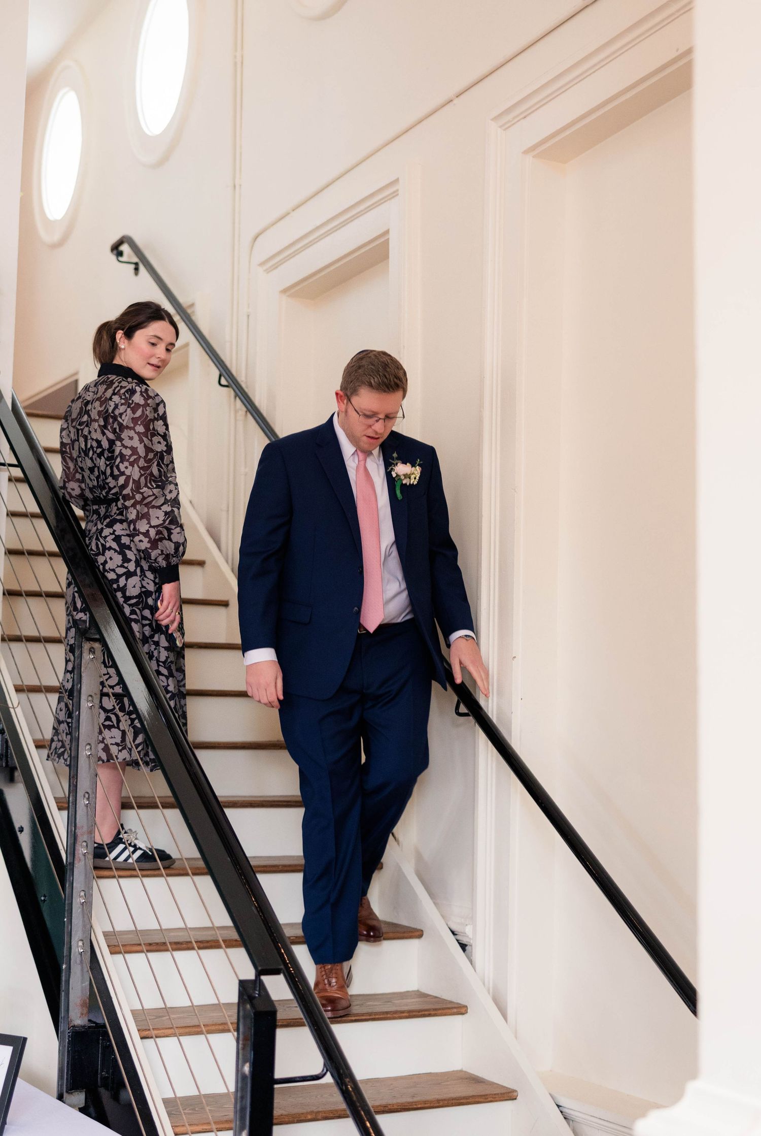 Groom in navy suit with boutonniere descends elegant staircase with female guest at wedding venue.