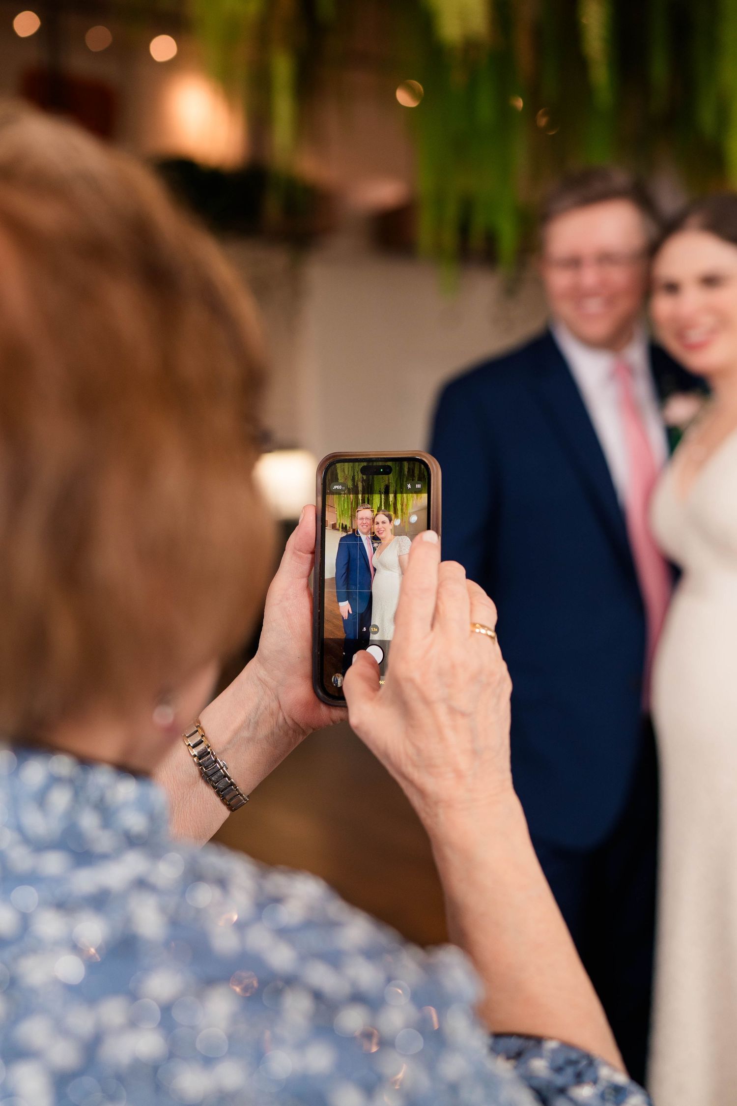 Woman photographs smiling wedding couple on smartphone at elegant indoor reception venue.