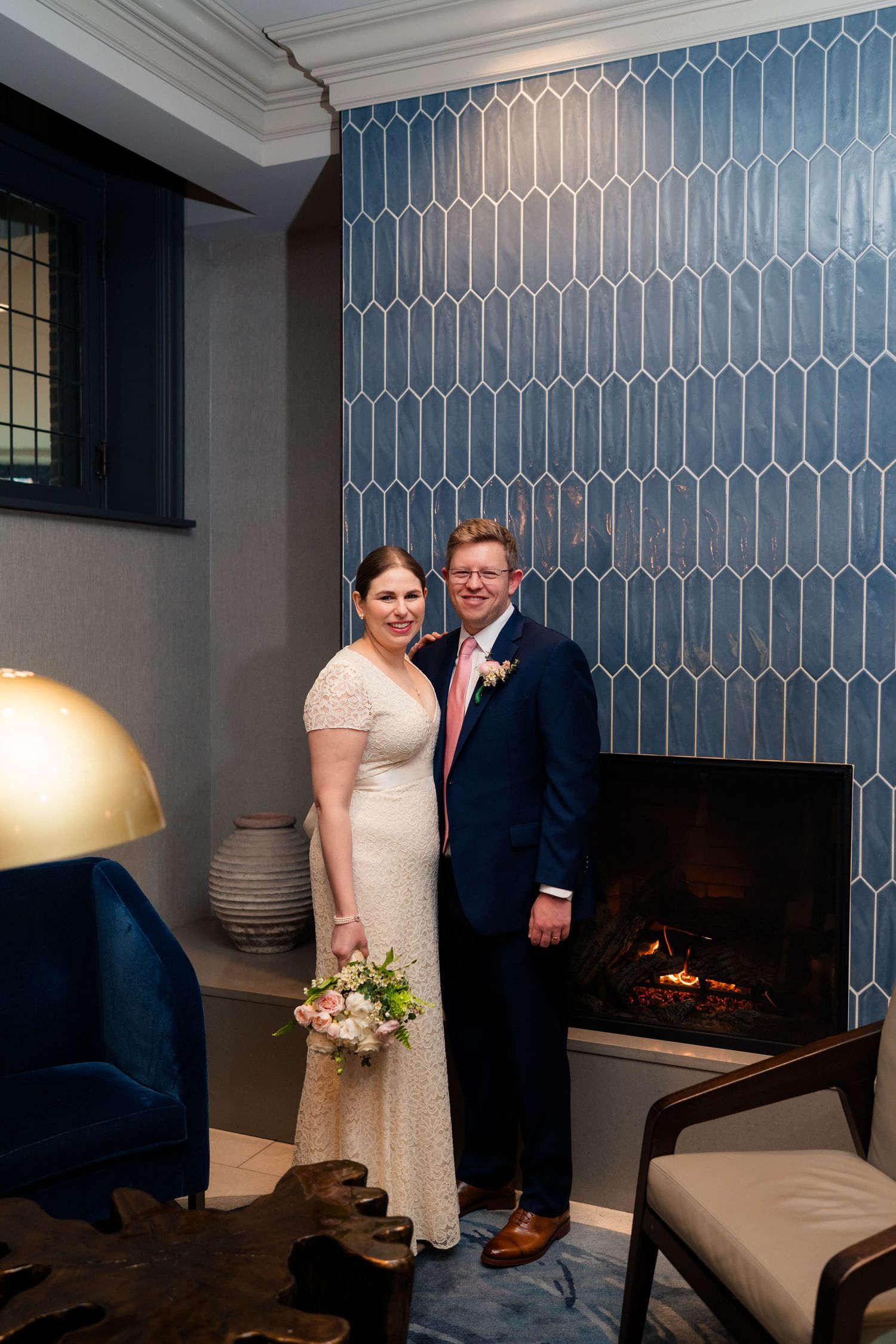 Bride and groom smiling together in front of a blue geometric tile fireplace, bride holding a pink floral bouquet.