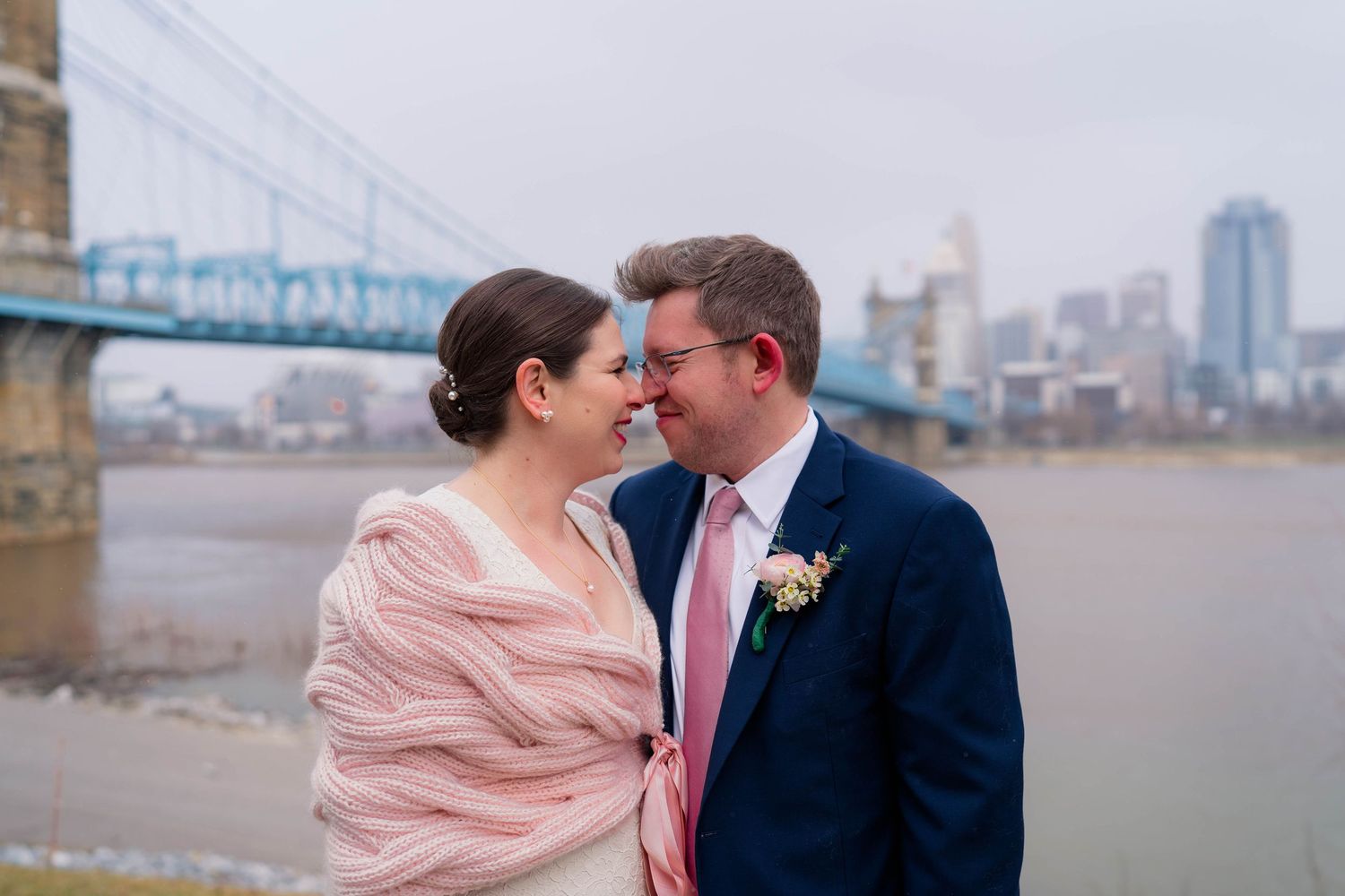 Smiling bride and groom sharing romantic moment with blue suspension bridge and river backdrop.