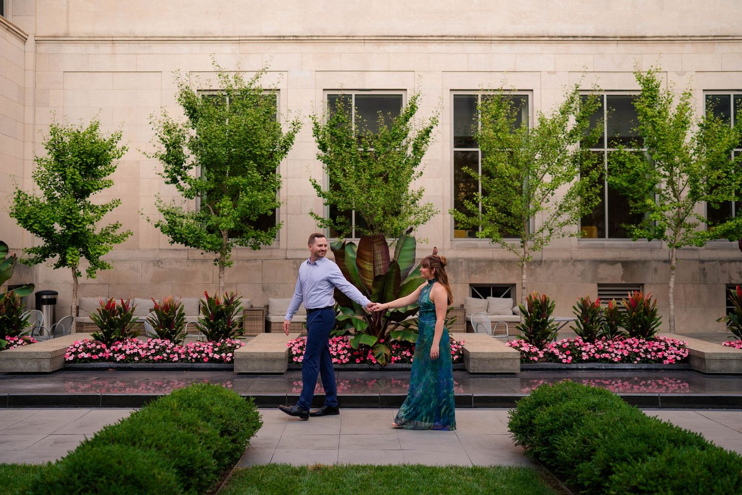 Couple holds hands in a garden courtyard with flowering beds and trees outside a stone building.