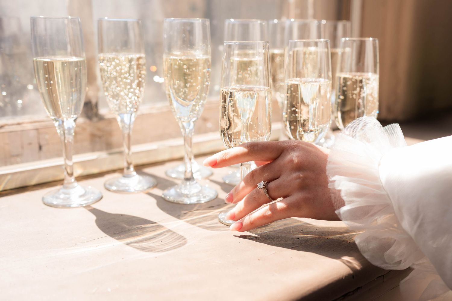 Bride's hand with engagement ring rests beside champagne flutes on sunlit windowsill.