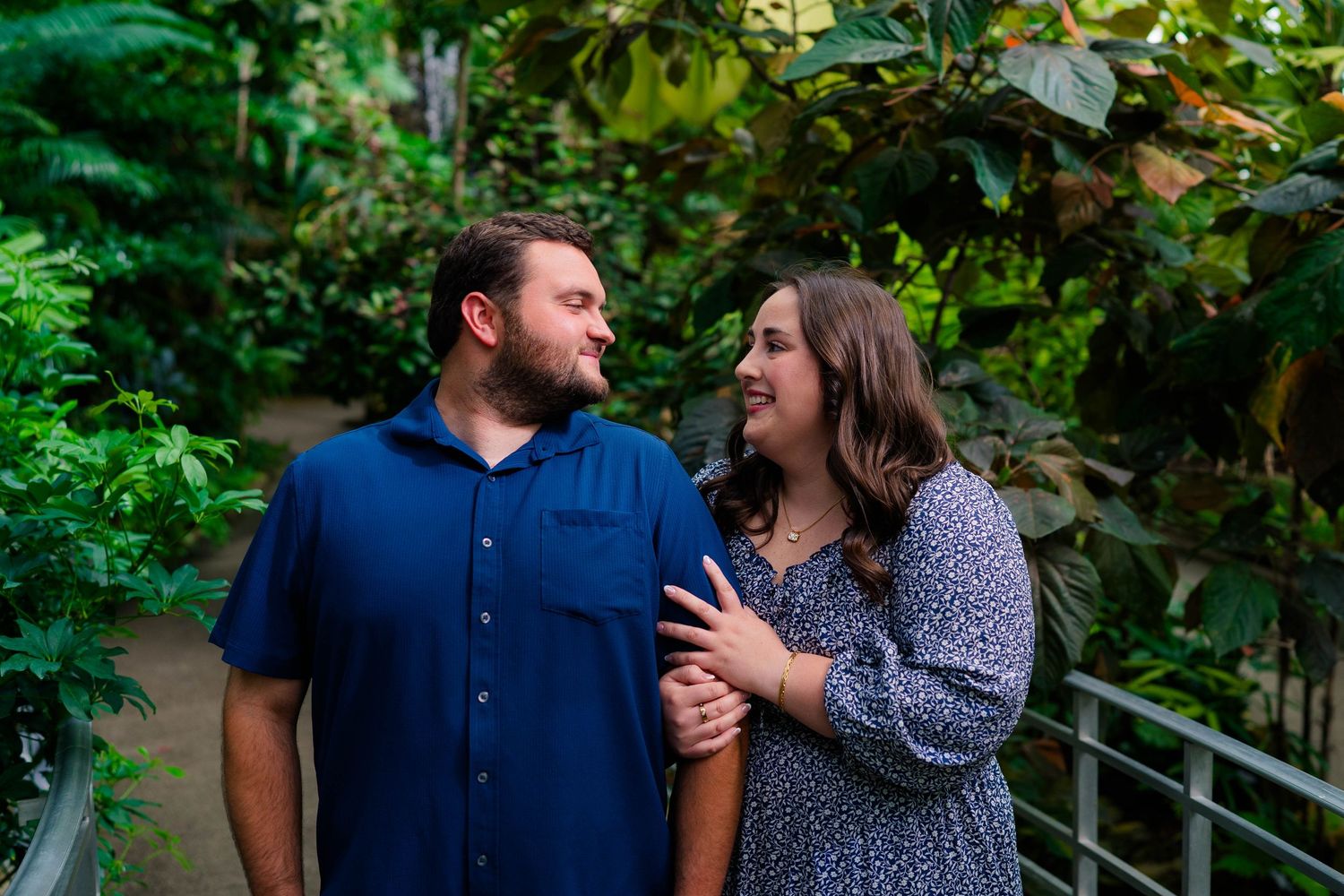 Couple smiles at each other while standing among lush tropical greenery in a botanical garden.