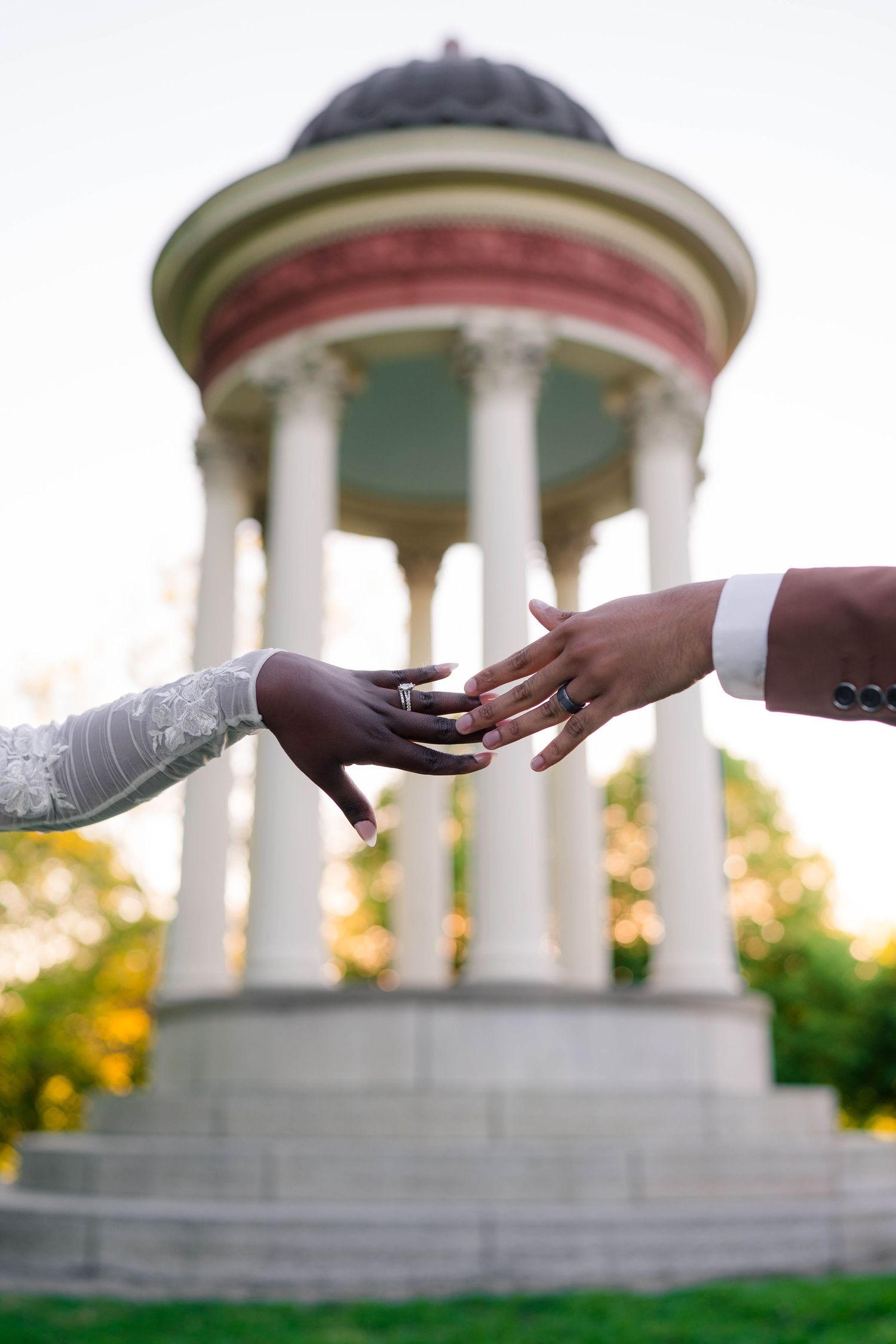 Close-up of Black bride and groom reaching hands toward each other with white columned gazebo behind.