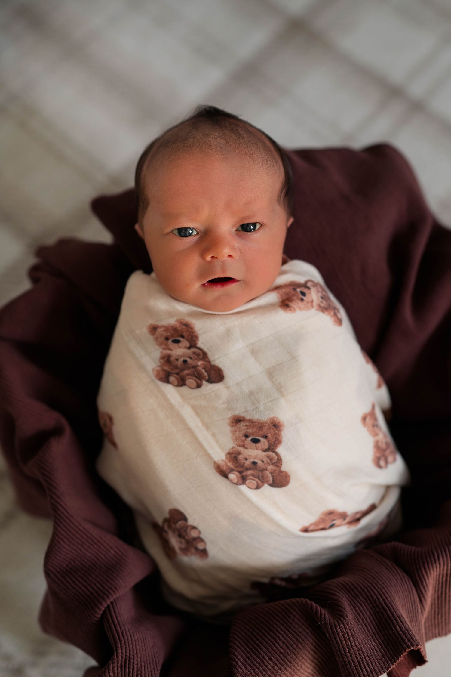 Newborn baby wrapped in white teddy bear muslin swaddle blanket resting in brown fabric bowl.