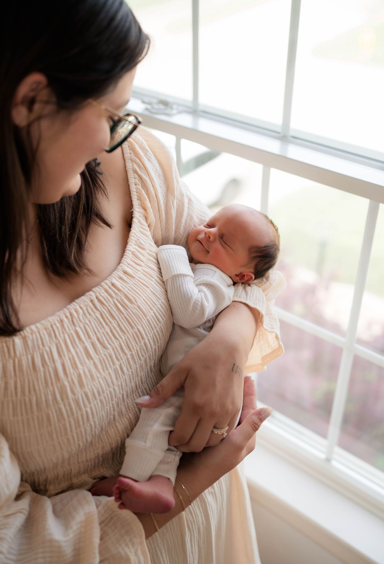 Mother in cream dress tenderly holds sleeping newborn baby near a bright window with soft natural light.
