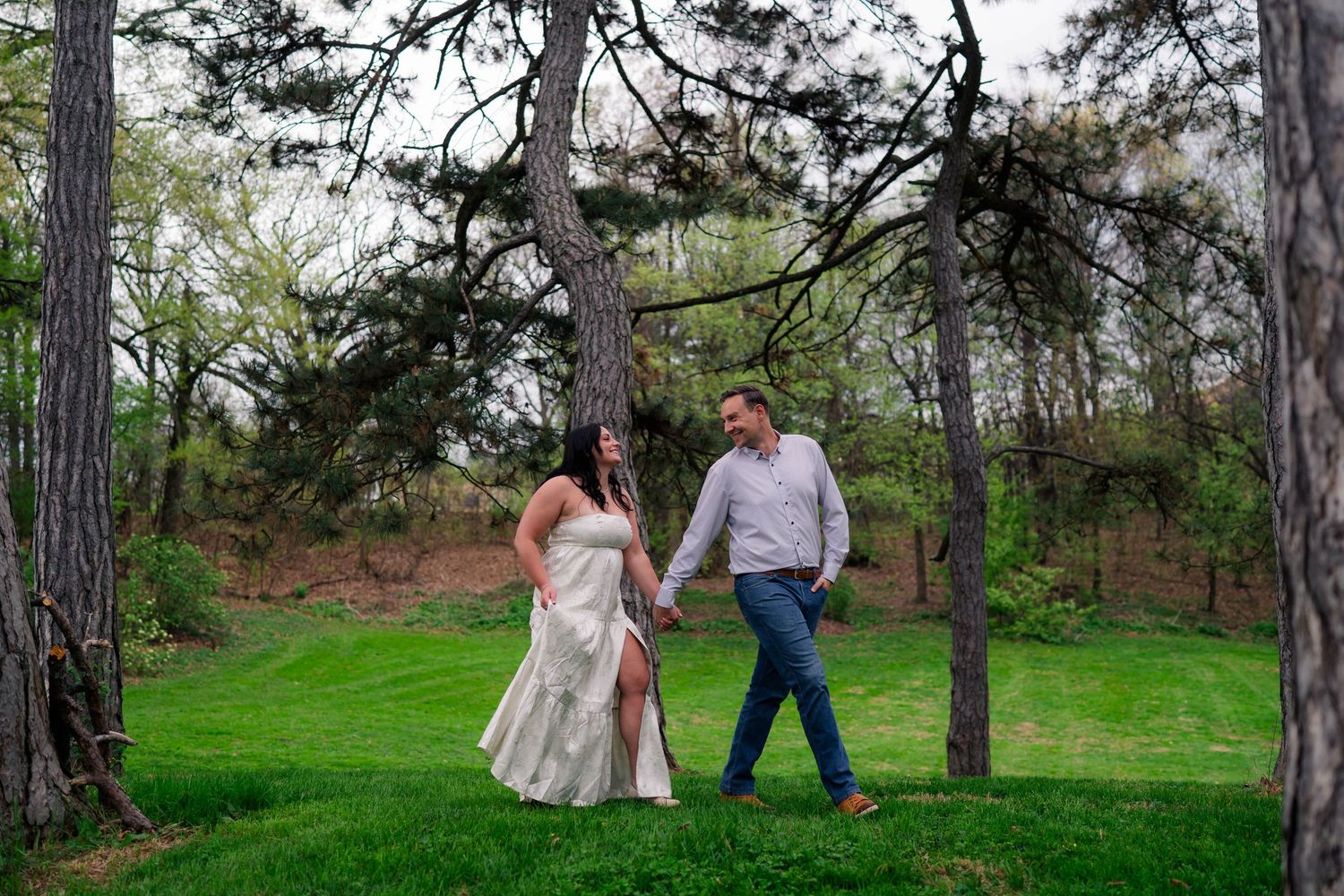 Engaged couple walking hand in hand through a scenic park with tall pine trees and vibrant green grass.