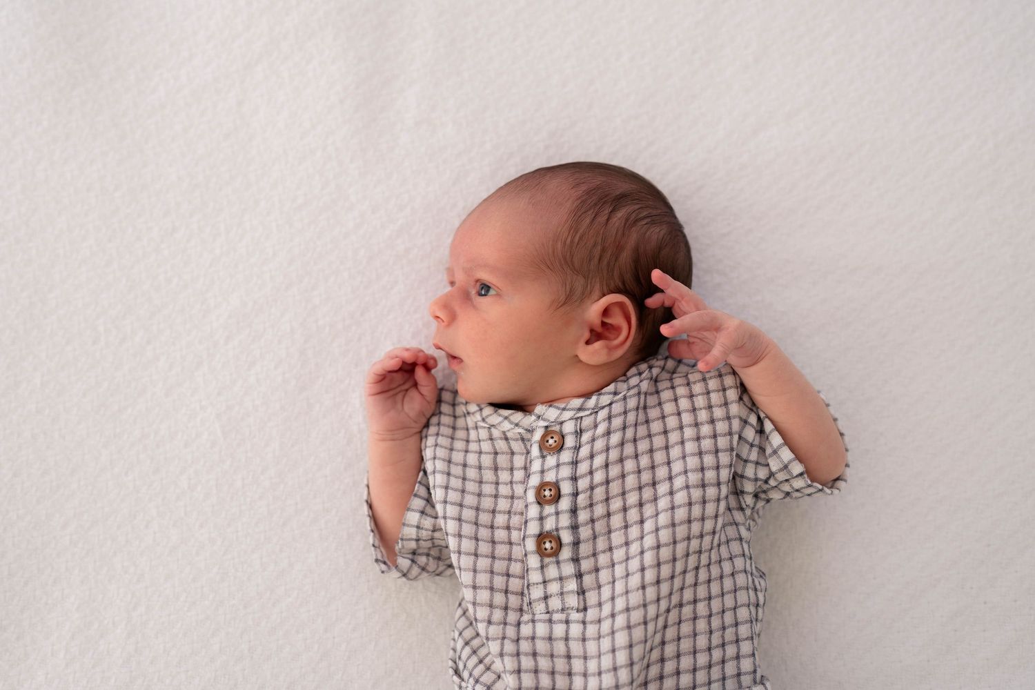 Newborn baby in a plaid button-up onesie lying awake on a white textured blanket.