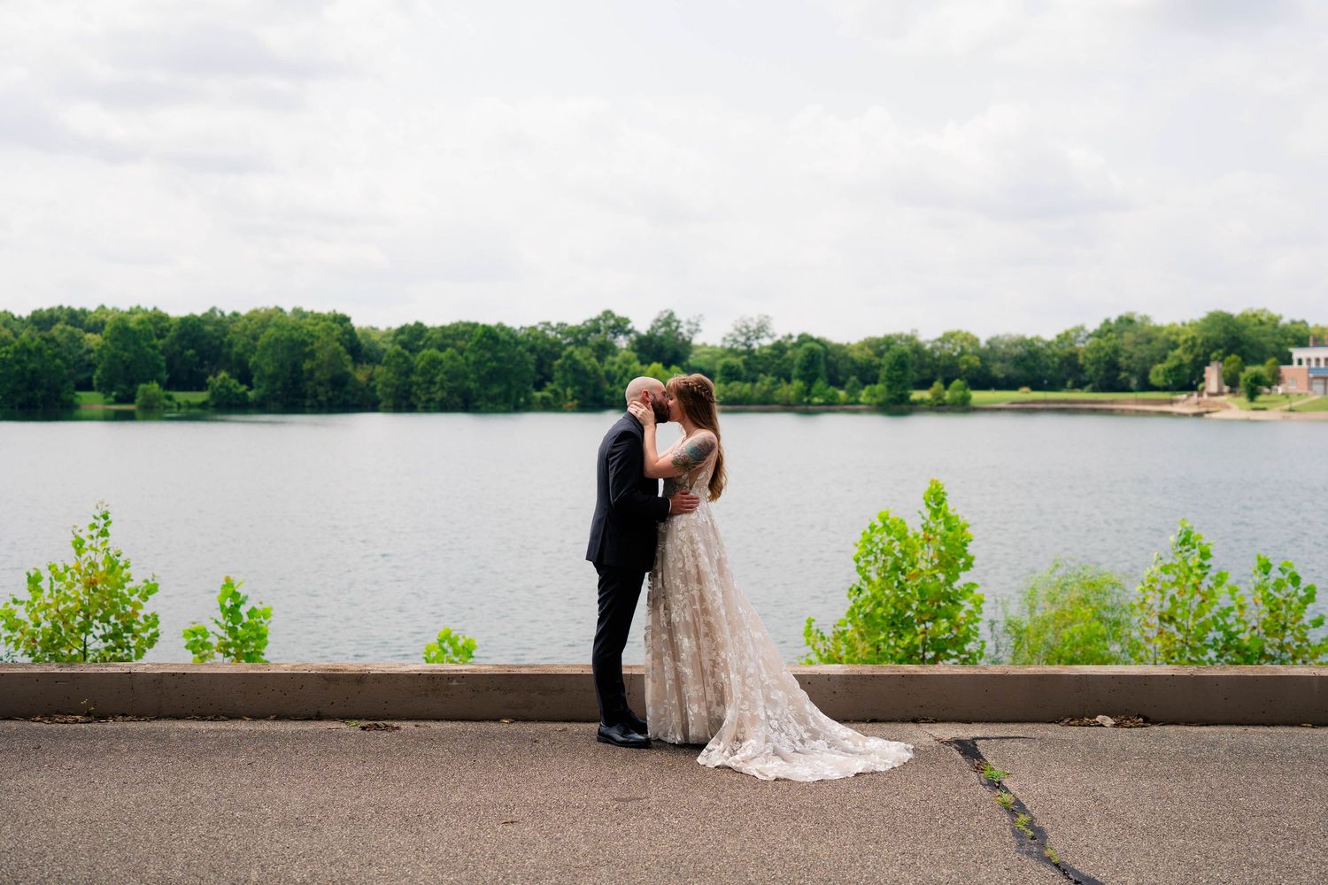 Bride and groom embrace romantically by a serene lake surrounded by lush greenery on their wedding day.