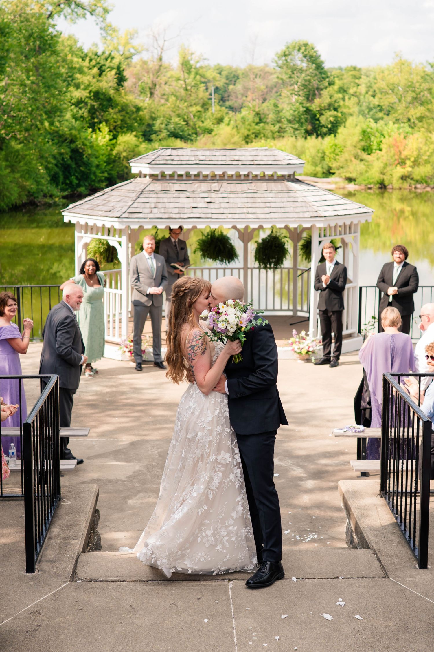 Newlyweds kiss at bottom of gazebo steps while guests celebrate with confetti by lakeside venue.