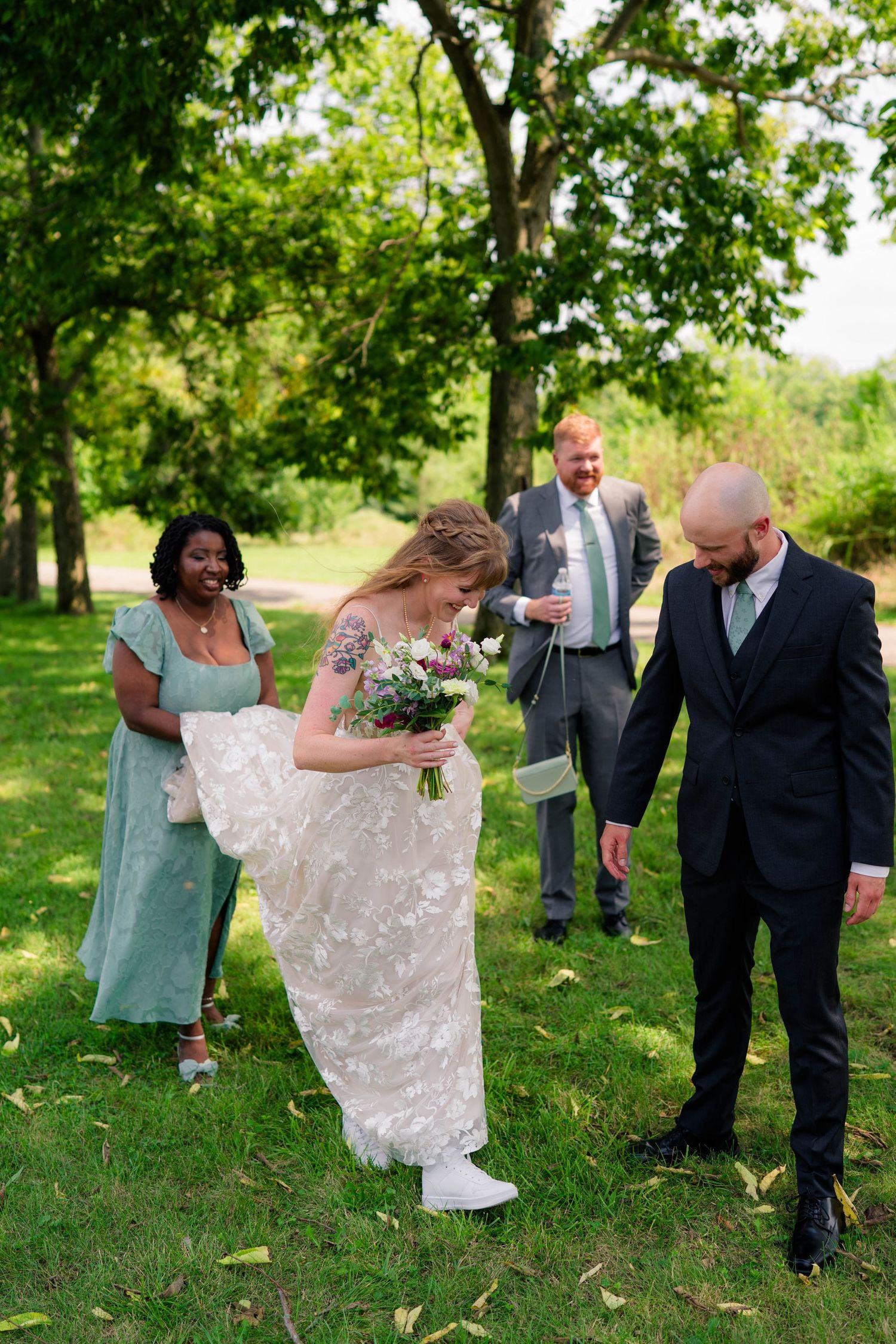 Bride in lace gown holds bouquet while groom in dark suit assists her walking on green lawn at outdoor wedding.