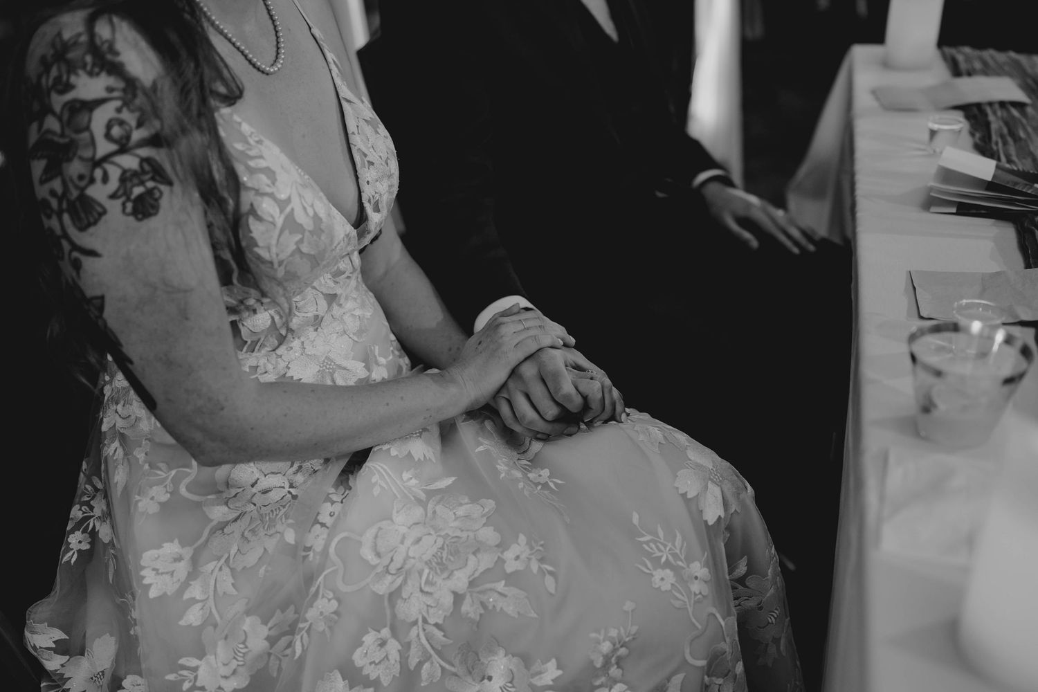 Black and white photo of bride in floral gown holding hands with groom at reception table.