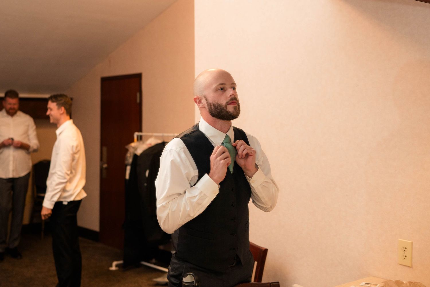 Groom adjusting green tie while wearing black vest and white dress shirt in hotel room before wedding.