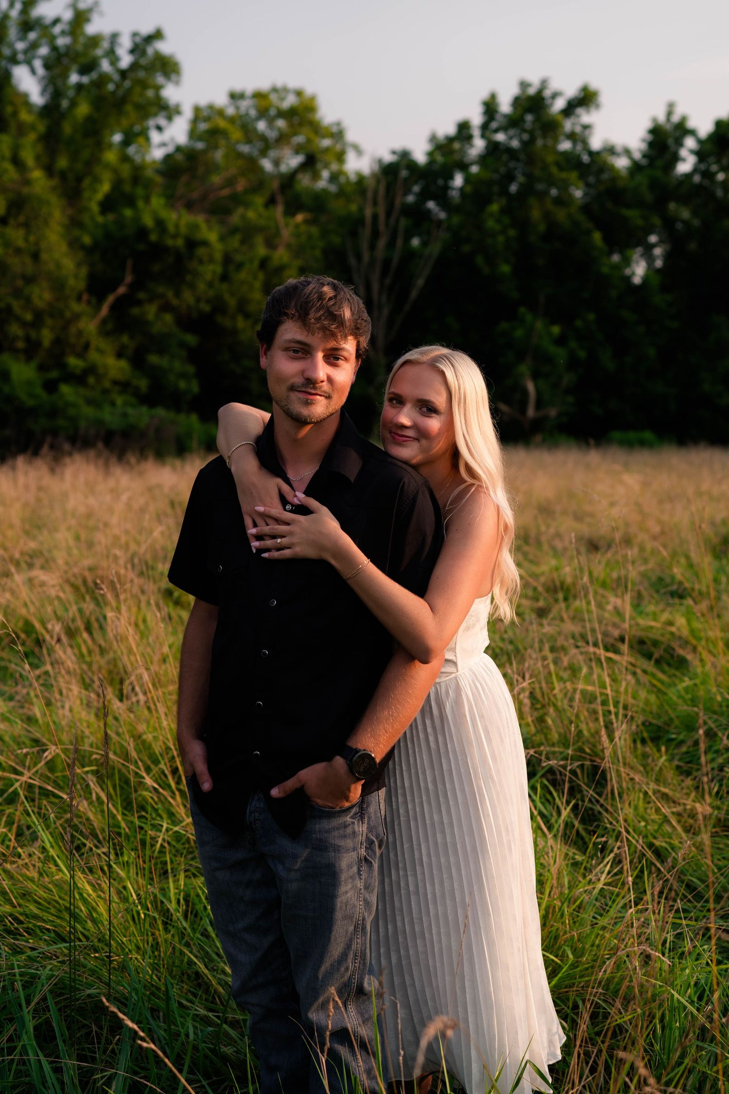 A young couple embraces in a golden field at sunset, woman in white skirt hugging man in black shirt.