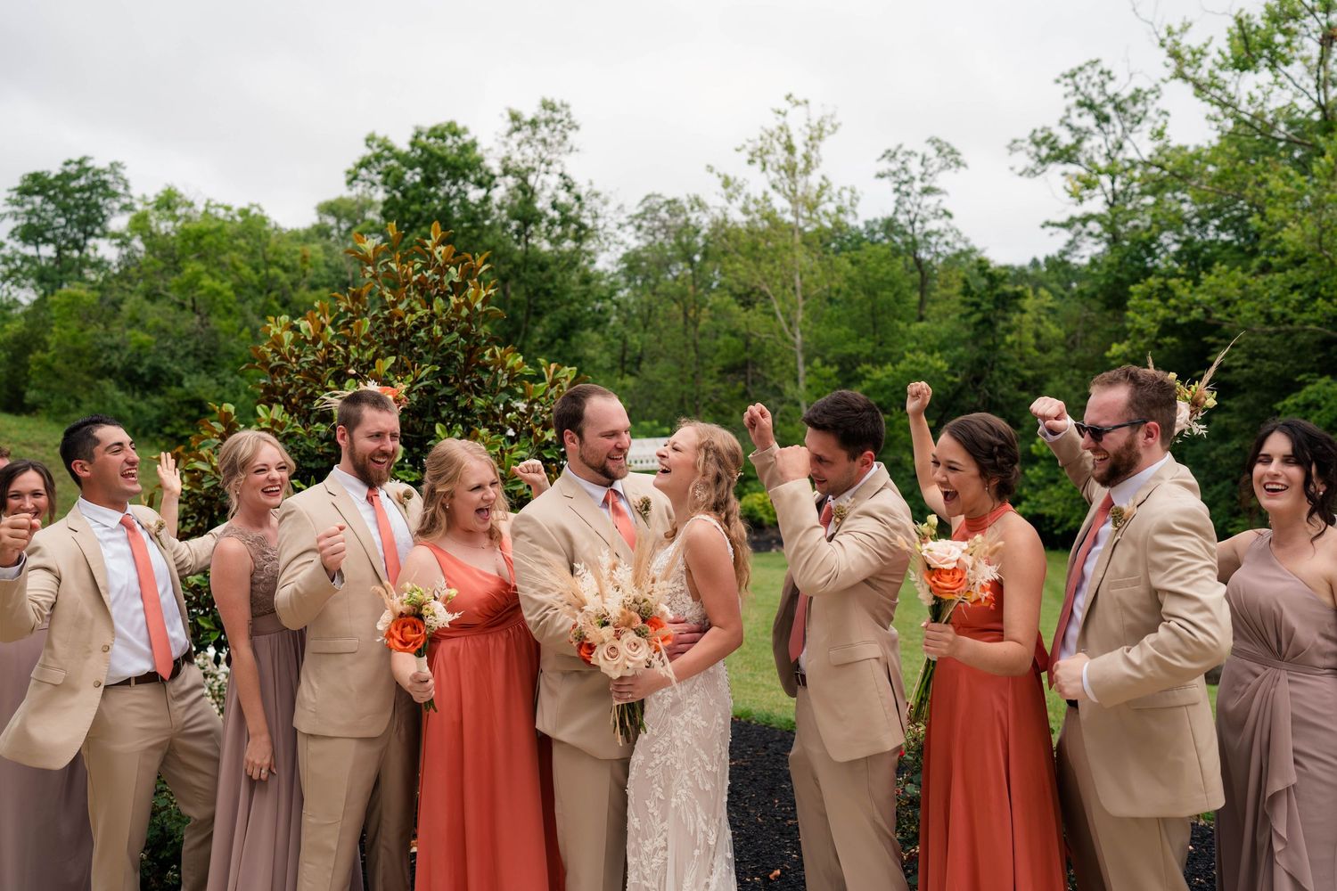 Wedding party in tan and coral attire celebrating joyfully outdoors in lush greenery.