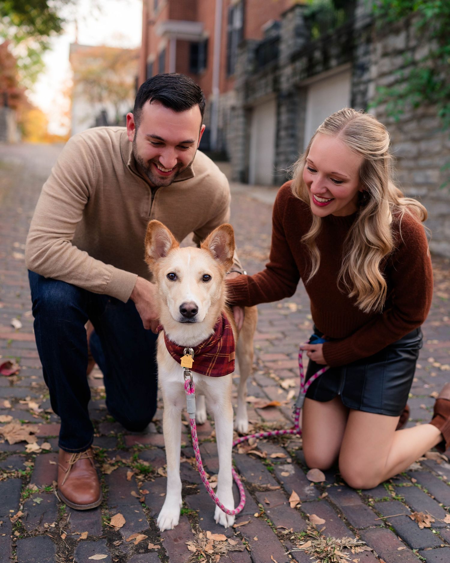Smiling couple kneeling on brick path petting their tan dog on a colorful autumn day.