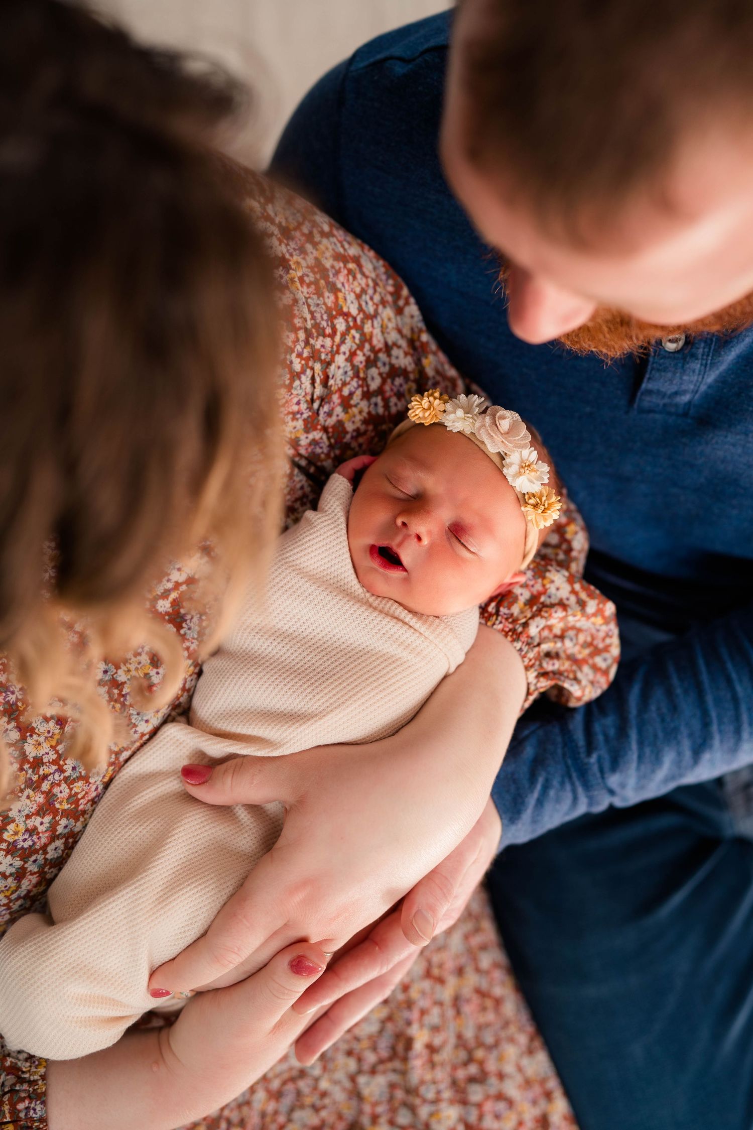 New parents in floral and blue outfits cradling sleeping newborn with yellow flower headband.