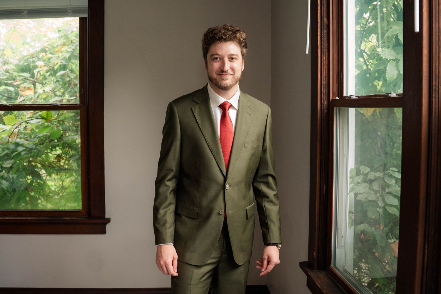 Man in olive green suit and red tie standing confidently between windows with green foliage backdrop.