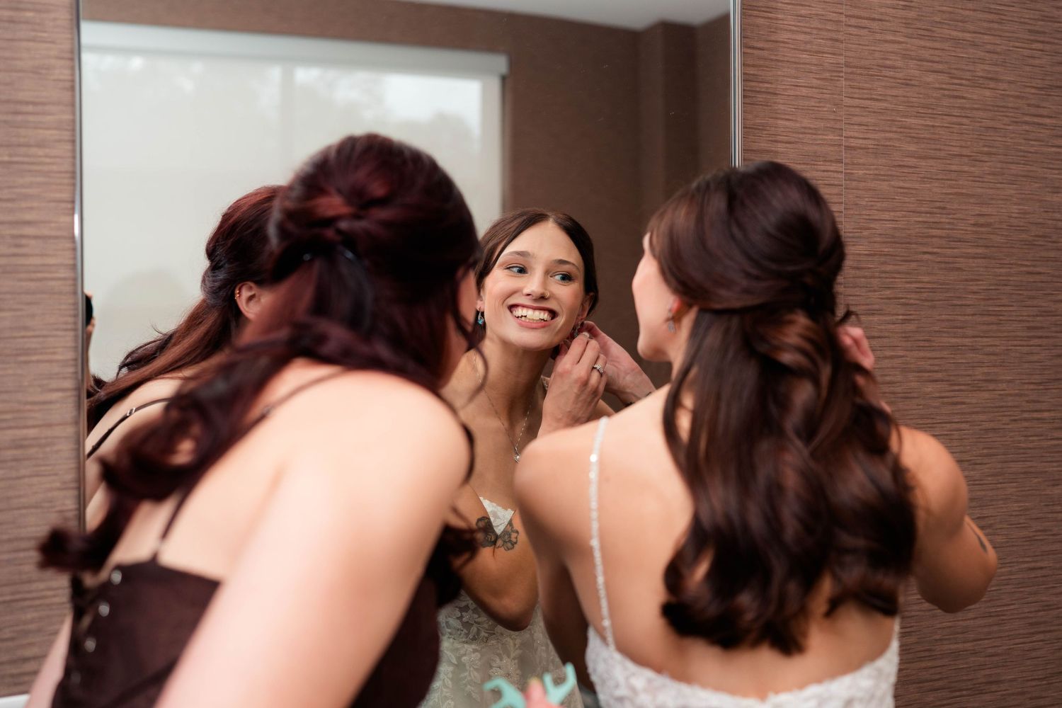 Bride smiling at reflection in mirror while bridesmaid helps with earring before wedding ceremony.