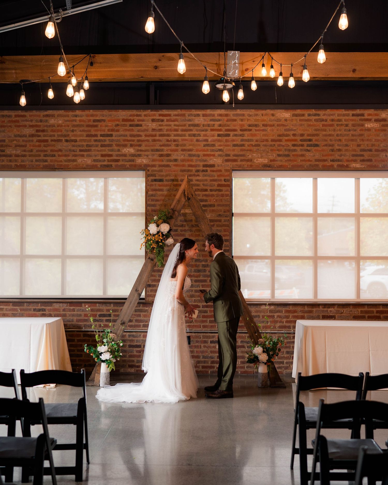 Bride and groom exchange vows at industrial wedding venue with exposed brick walls and Edison bulb lighting.
