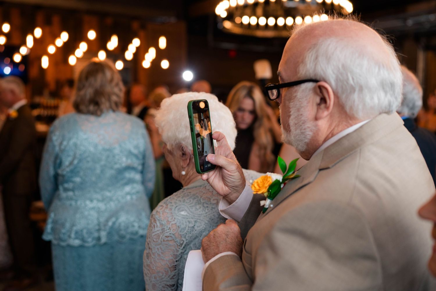 Elderly wedding guest photographs the celebration on a smartphone in a warmly lit reception venue.
