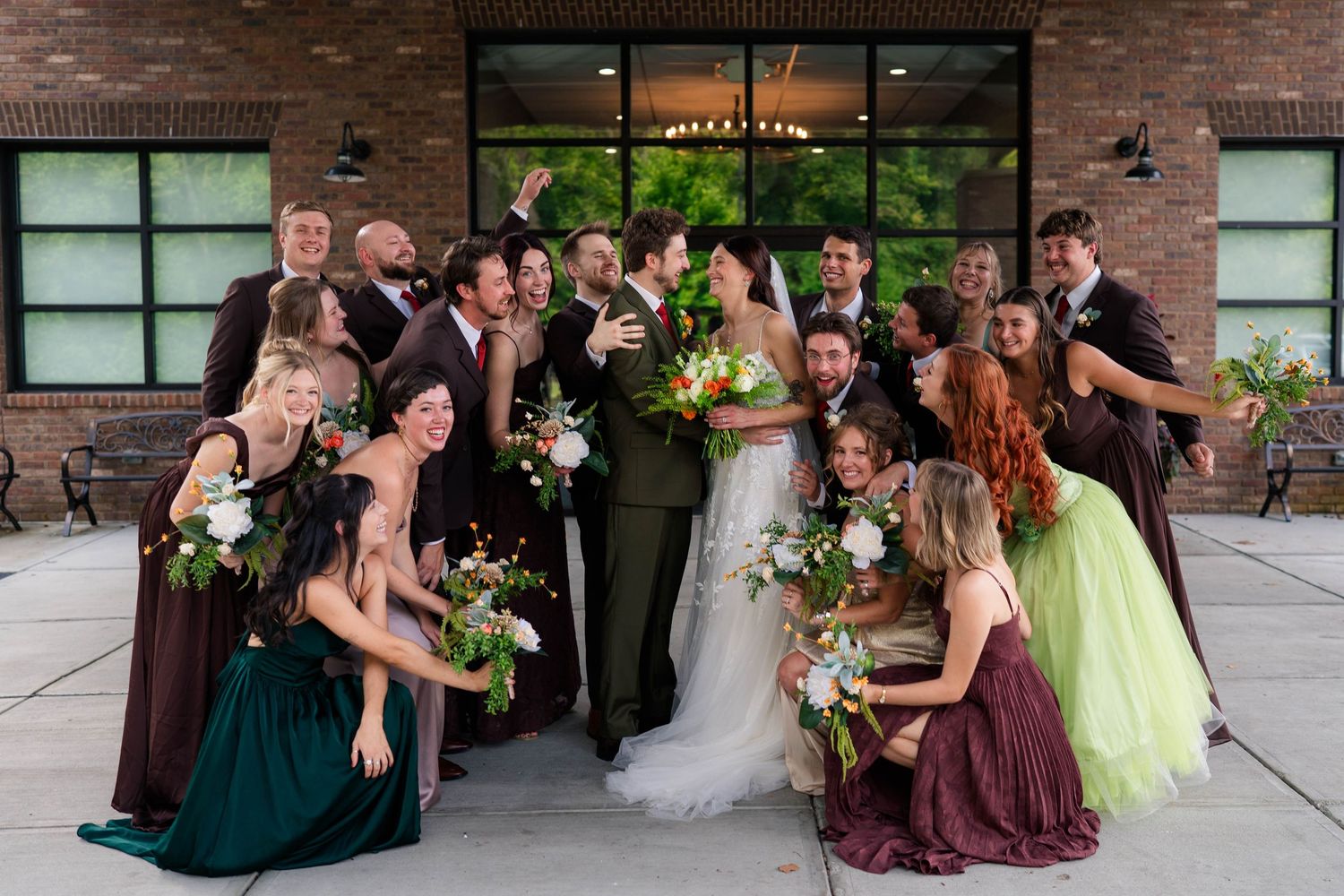 Large wedding party celebrates together outside a brick venue, bridesmaids in green and burgundy gowns.