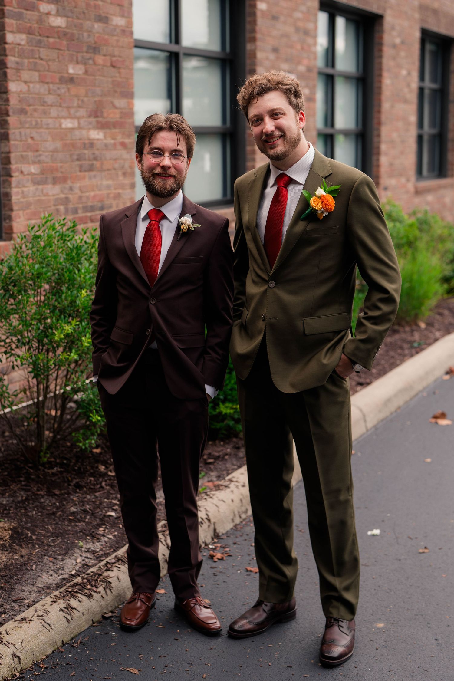 Two groomsmen wearing dark suits with red ties and floral boutonnieres stand outside brick building.