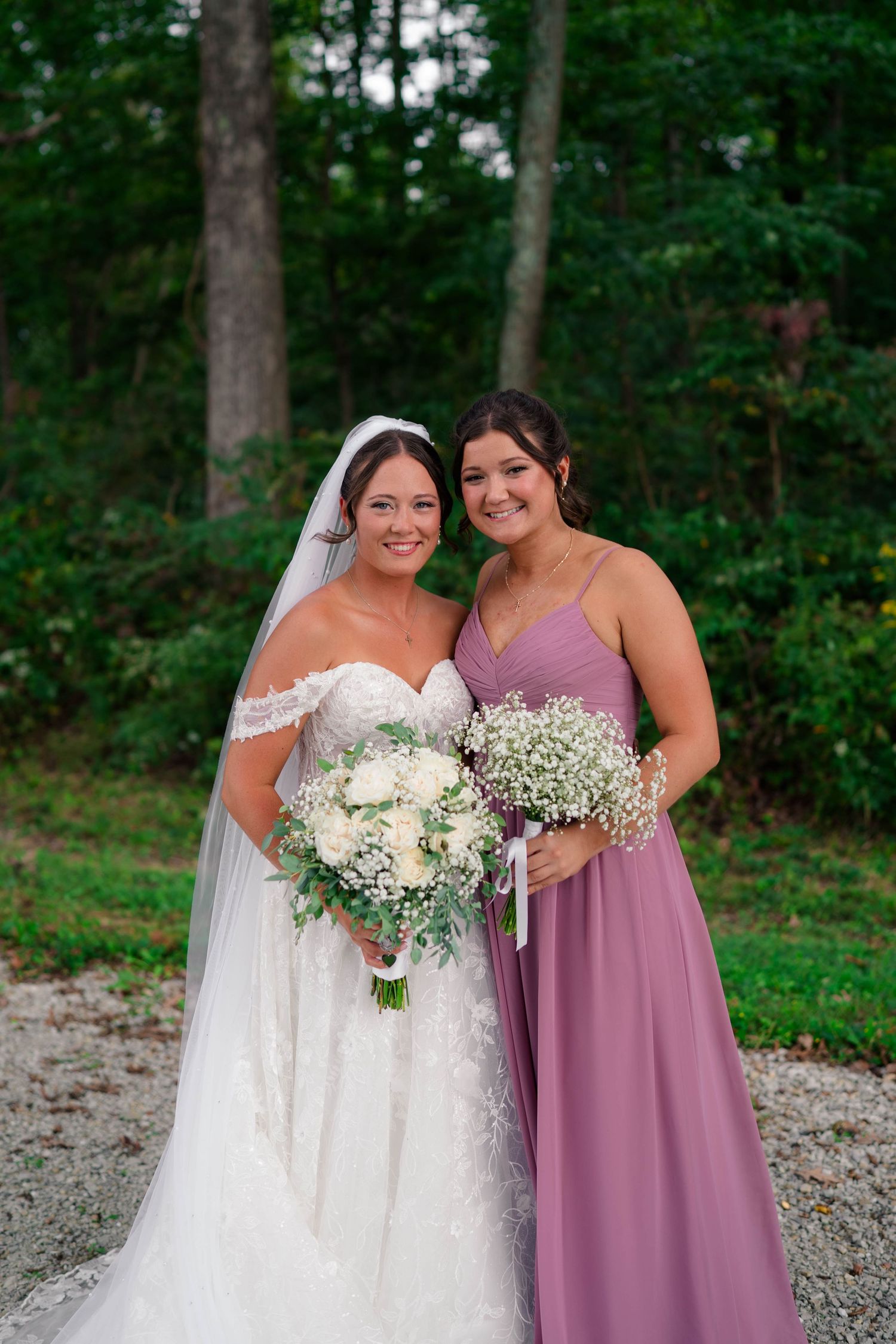 Bride in white lace gown and bridesmaid in mauve dress holding bouquets outdoors near trees.