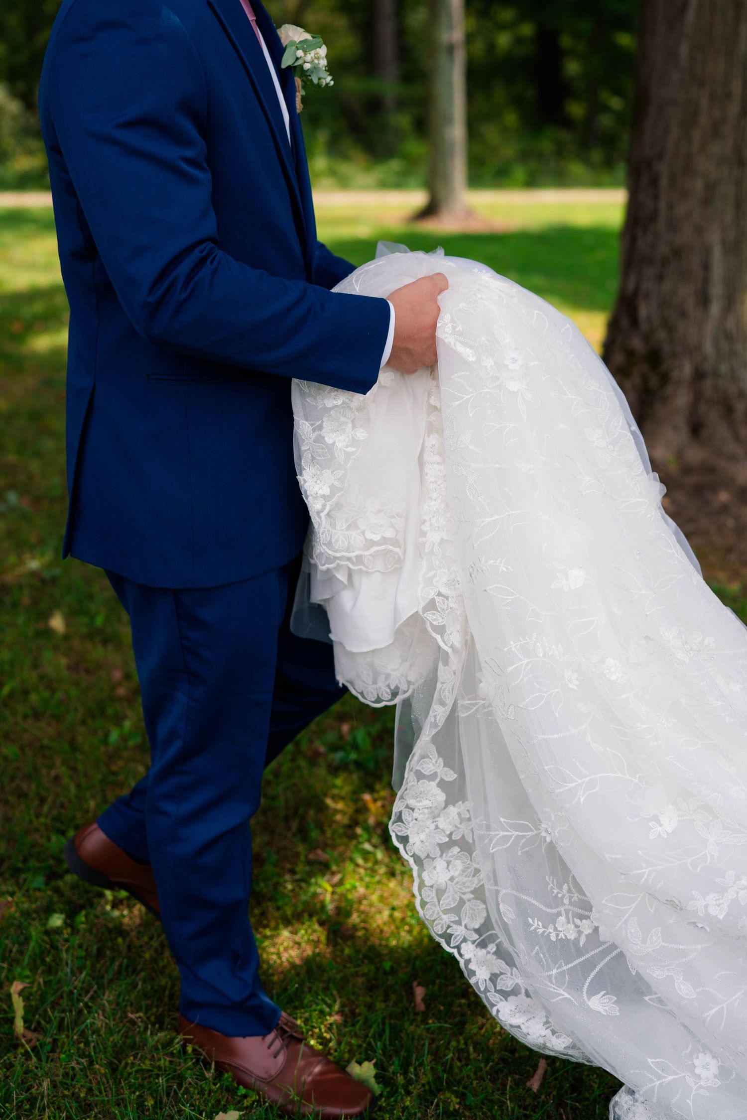 Groom in navy suit gallantly carrying bride's white lace wedding gown train outdoors.