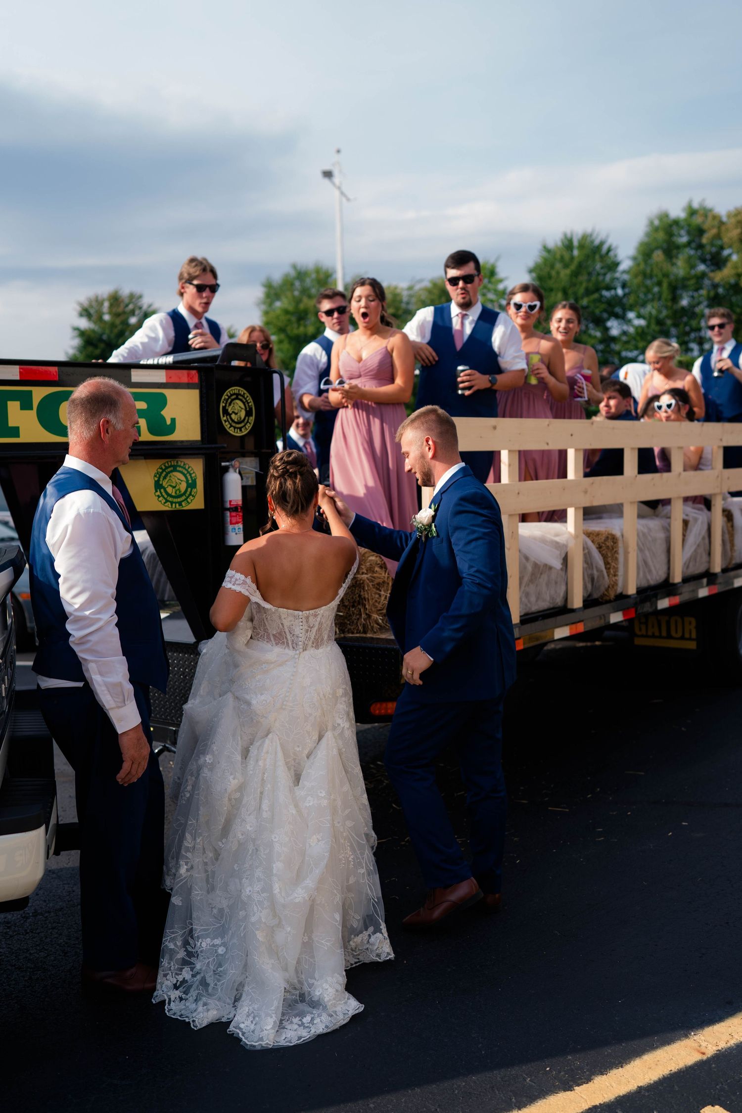 Bride and groom boarding a decorated flatbed trailer with wedding party at outdoor celebration.